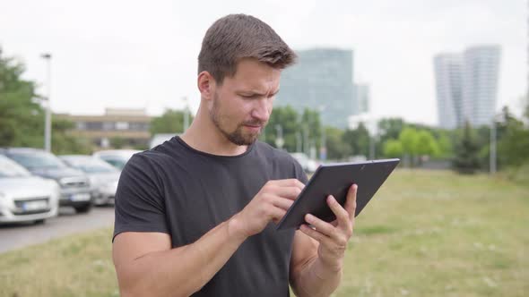 A Young Handsome Man Works on a Tablet By a Parking Lot - a City in the Background