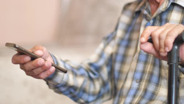 in the hands of a sitting pensioner a walking stick and a smartphone. an elderly man uses the phone.