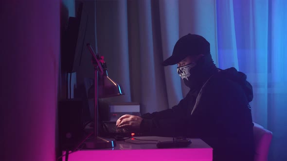 Male Hacker Sits at His Desk and Breaks Into the System. Man in Black Hoodie with Bandana and Black