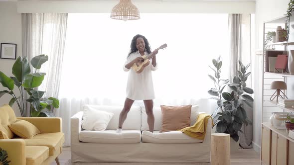 Motivated Woman Playing Ukelele Standing and Jumping on the Sofa Living Room