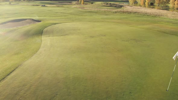 Moving over a green beautiful golf course with a white flag close to a sand bunker.