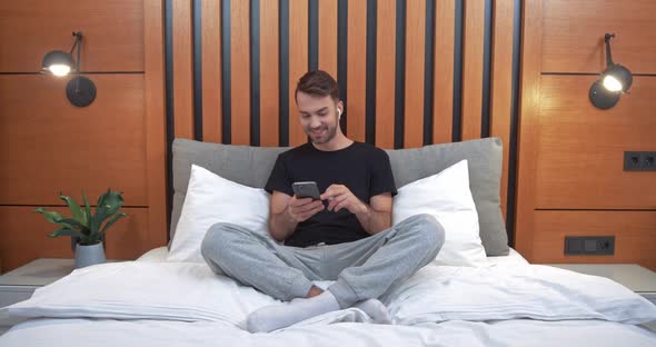 Young Happy Man Sitting on the Bed in the Bedroom and Listening To Music in Wireless Headphones