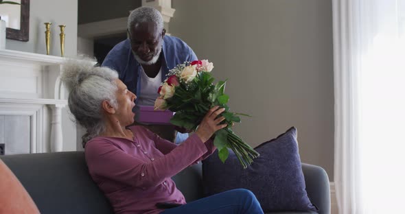 African american senior man giving gift box and flower bouquet to his wife at home