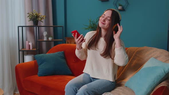 Overjoyed Young Woman in Wireless Headphones Dancing Singing on Cozy Couch in Living Room at Home