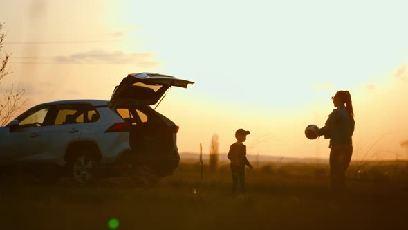 Mom and Baby Son in Nature Playing Ball in Field with Tall Grass Against the Back of Sunset and Car