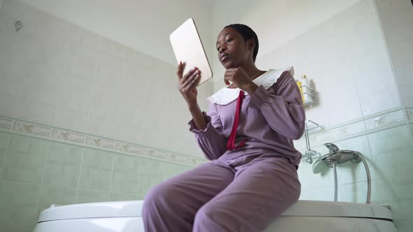 Concentrated Young African American Woman Looking in Hand Mirror Sitting in Bathroom on Bathtub