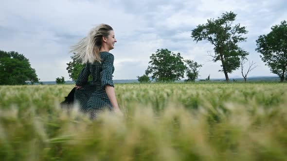Young Happy Blonde Girl Runs on a Green Wheat Field in the Evening Against the Background of the