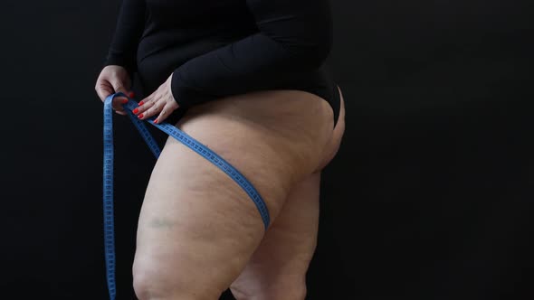 a Fat Woman with Cellulite Measures Herself with a Measuring Tape on a Black Background