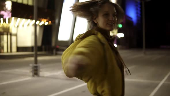 Cute Cheerful Girl Whirls Around Herself on a Lit Parking Place at Night