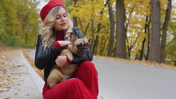 Beautiful Blonde Woman in a Red Suit Holding a French Bulldog in Her Arms