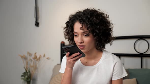 Smiling Hispanic Curly Woman Uses Smartphone Dictating Audio Message to Friend
