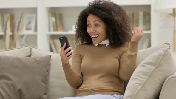 Young African Woman Celebrating Success on Smartphone on Sofa