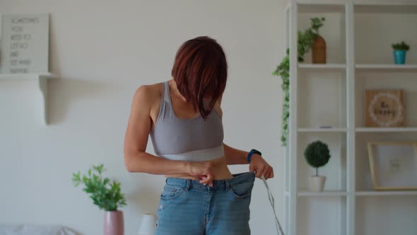 Young woman admiring the result of weight loss while wearing old jeans in bedroom.