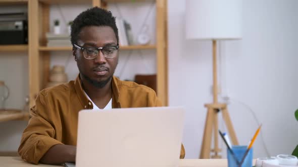 African Young Man in Glasses Works on Laptop Against Background of Home Interior Spbas