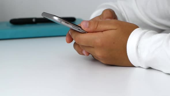 A child girl holds a phone in her hands, sits at the table, and does her homework. Close-up video.