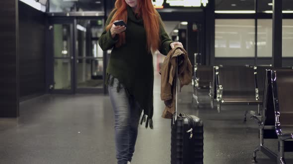 Young Woman Walks Through an Airport Terminal and Uses Smartphone