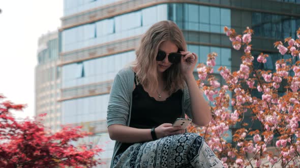 Closeup of Beautiful Young European Girl in Sunglasses Sitting on the Street and Using Smartphone