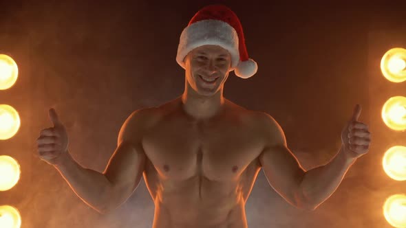 Portrait of Muscular Man Wearing Christmas Santa Hat Showing Thumb Up on Smoky Background