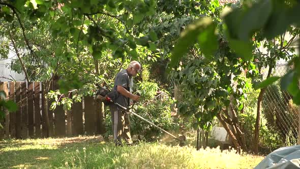 Man Trimming Overgrown Grass In The Backyard With Grass Cutter. wide