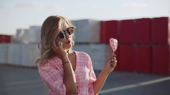 Beautiful Young Woman Uses a Smartphone on the Street and Licks Colored Lollipop