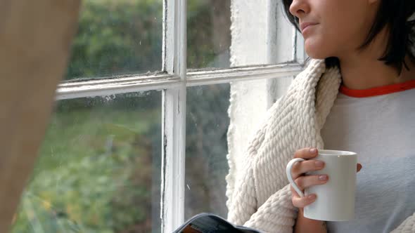 Pensive woman sitting on windowsill and having cup of coffee