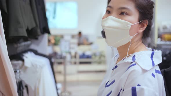 asian female shopper wearing facial virus protective mask choosing clothes at the store