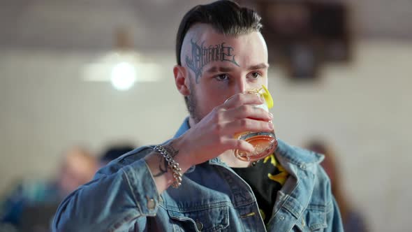 Handsome Confident Young Man with Tattoo on Face and Piercing Drinking Cocktail in Slow Motion