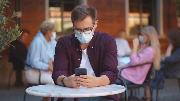 Young Guy Wearing Facial Mask Surfing Internet on Mobile Phone in Outdoors Cafe