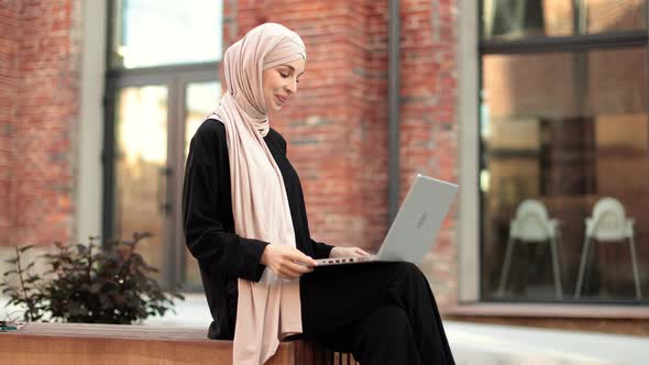 Muslim Woman in Hijab and Black Clothes Sitting on Bench During Break Talking on Laptop