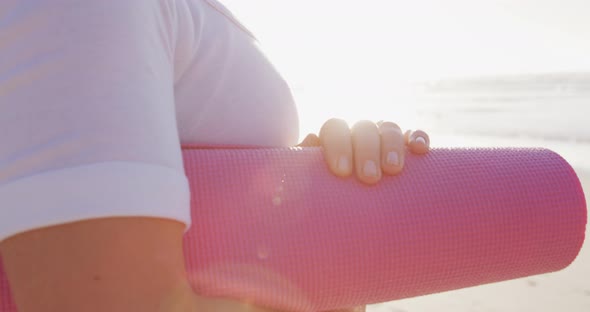 Caucasian woman holding pink sport mat on the beach and blue sky background