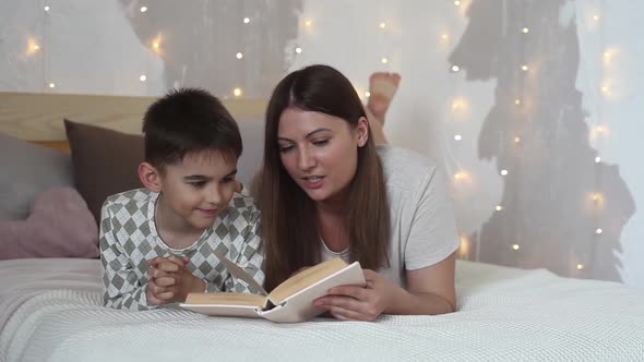 Beautiful Girl Reads a Book to a Small Boy Lying on a Bed and Kisses Him