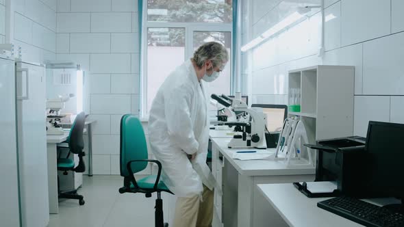 Laboratory Assistant Sits on a Chair and Looks Through a Microscope in White Lab