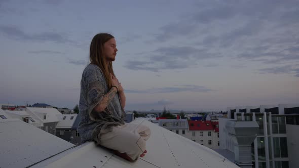 Young Man With Long Hair Meditating On City Roof