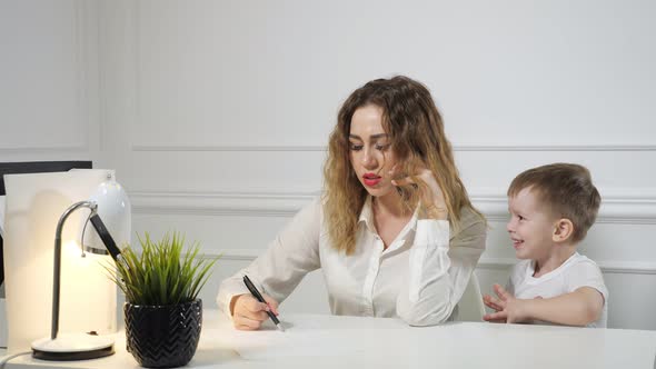 Little Boy is Stopping His Mom to Work While She is Calling Phone in Her Workplace in Office