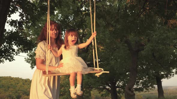 Mom Shakes Her Daughter on Swing Under Tree in Sun