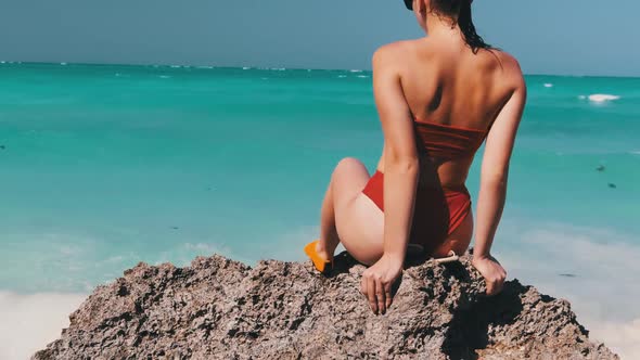 Young Woman in Red Bikini Sitting on Stone Reef and Looking at Ocean Rear View