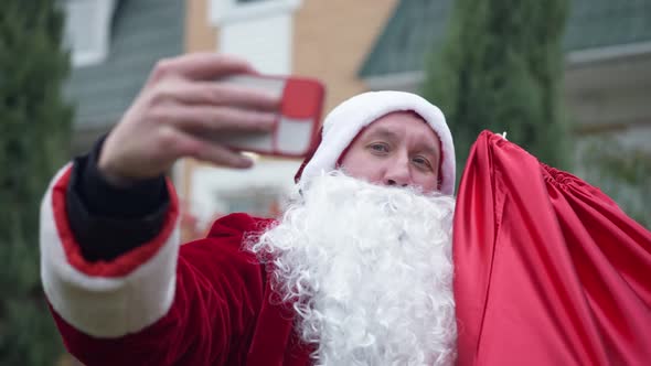 Caucasian Young Man in Red Santa Clause Costume Taking Selfie on Smartphone Standing Outdoors