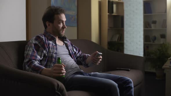 Guy Sitting on Sofa with Beer Bottle in Hand, Using Remote Control to Switch Tv