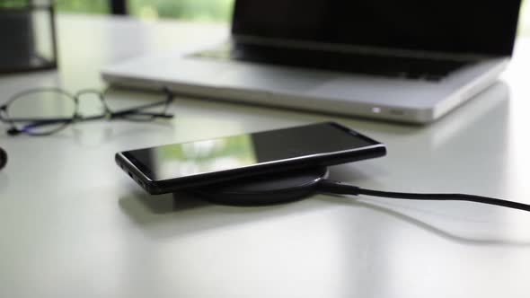 Smart Phone Being Charged on Wireless Charger Alongside with Laptop Glasses and Notebook on Table