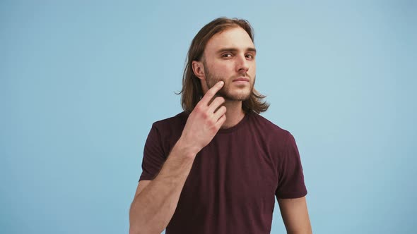 Young Man is Scratching His Beard Straightening Hair and Smiling Posing on Blue Background