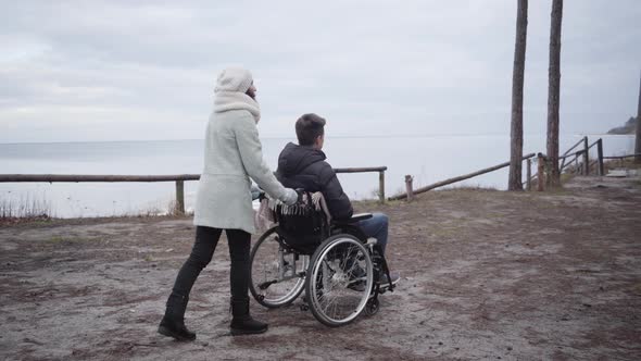 Back Side View of Woman Rolling Wheelchair with Young Caucasian Man Along Autumn River Bank. Invalid