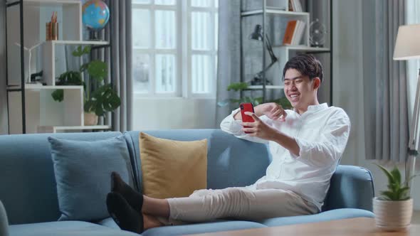 Happy Asian Man Having Video Call On Smartphone While Lying On Sofa In The Living Room