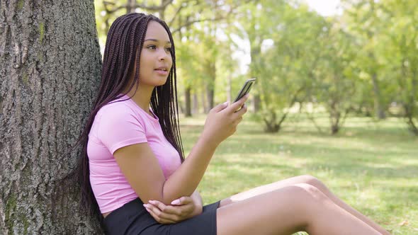 A Young Black Woman Works on a Smartphone with a Smile As She Sits Under a Tree in a Park