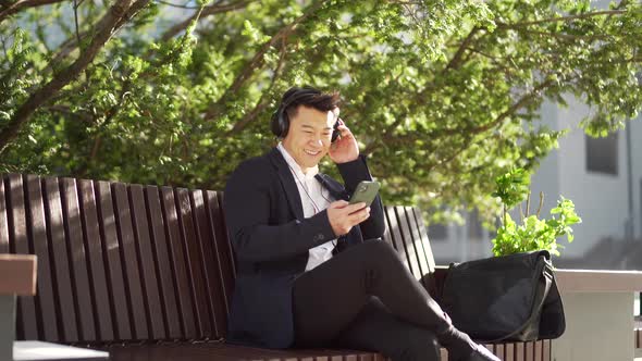 cheerful asian business man sitting on a bench in a city park in downtown and listening to music