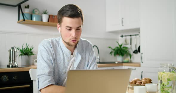 Man in Wireless Earphones Talks Online By Video Call Using Laptop in Kitchen