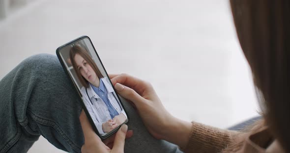 Female Medical Assistant Wears White Coat, Headset Video Calling Distant Patient on Smartphone