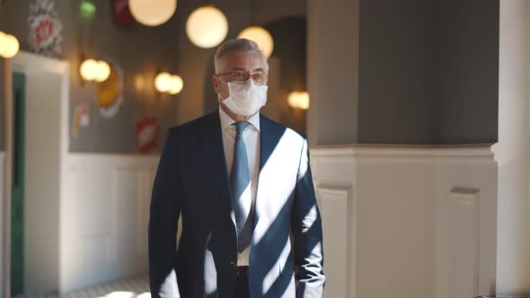 Senior Confident Businessman in Protective Mask Walking in Modern Office Corridor