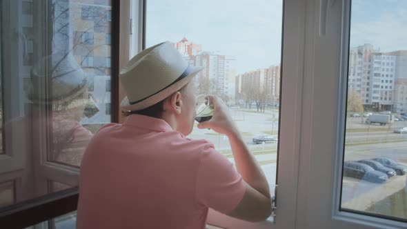 A Man in a Hat Stands on the Balcony Looking at the Street Through an Open Window and Drinks