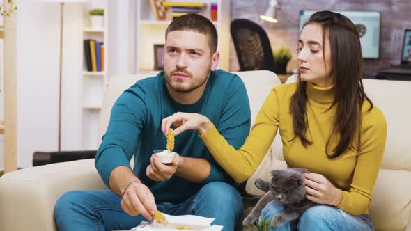 Concentrated Young Couple Sitting on Couch Eating Fried Chicken