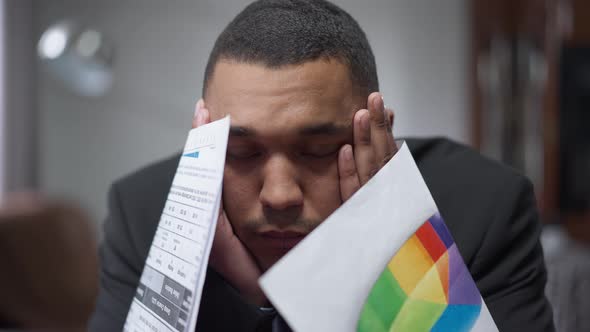 Closeup of Overwhelmed Exhausted African American Man Sighing Holding Graphs and Diagrams Sitting in